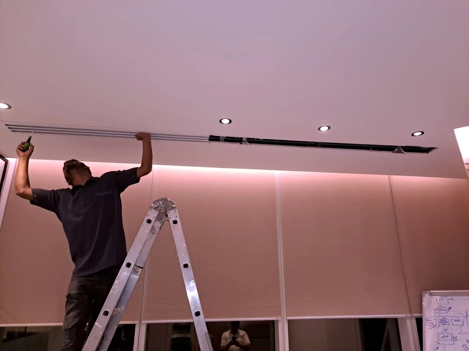 Technician on a stepladder fitting a linear air diffuser into a ceiling in a pink-lit room